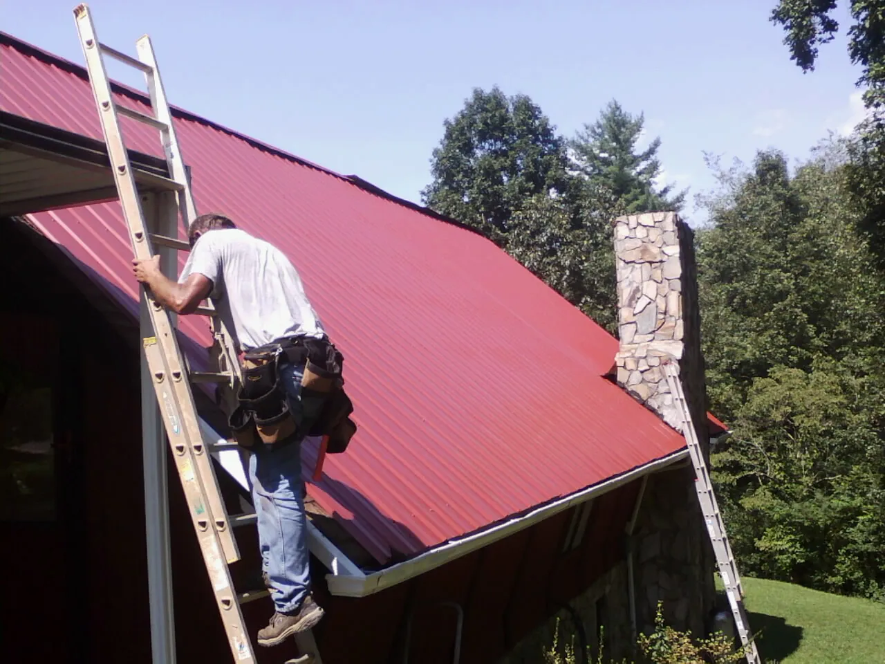 Roofer on a ladder beside a red metal roof and stone chimney — Soffit & Fascia Repair by Roofing Brothers in Moyock